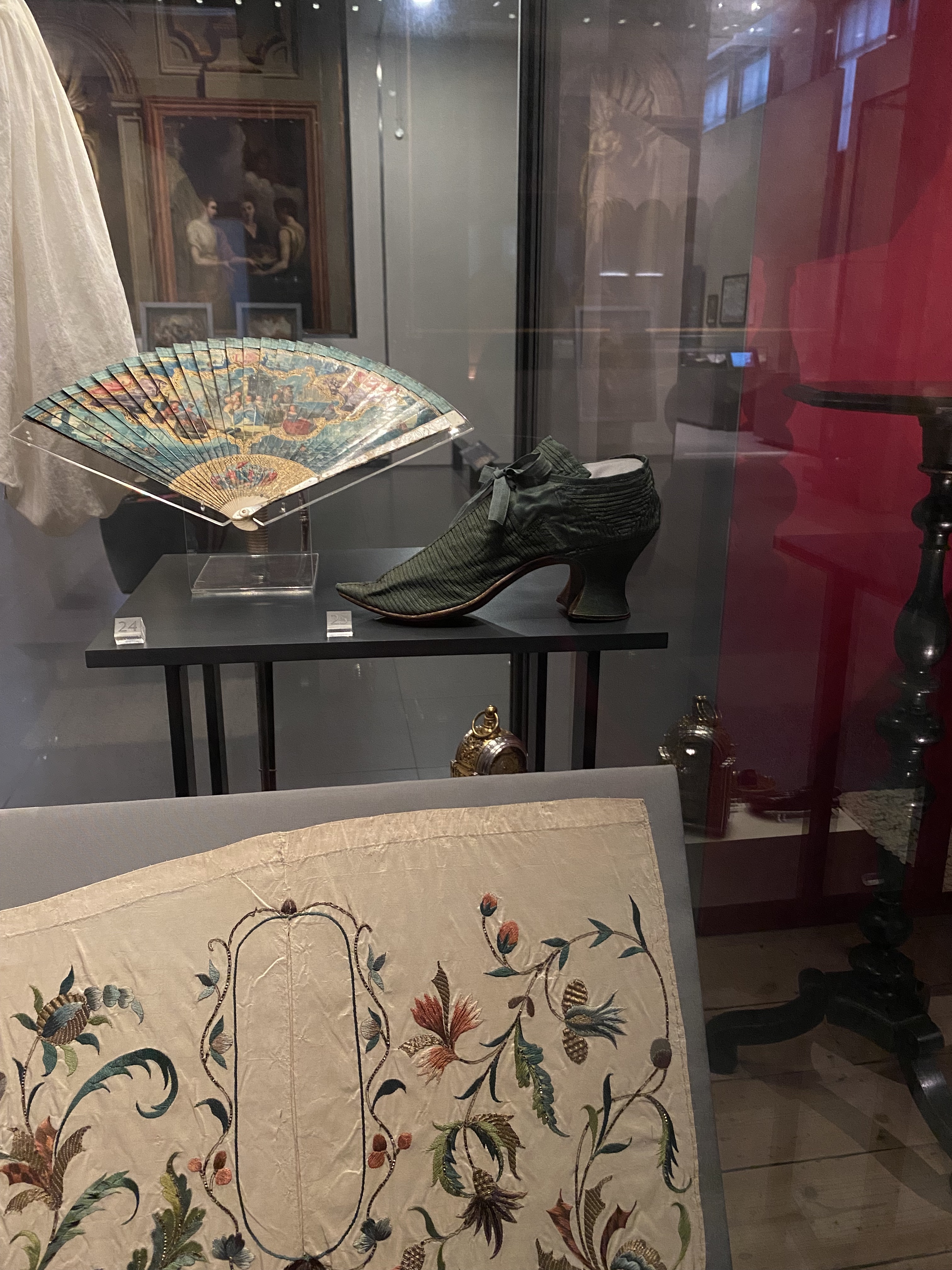a view inside a museum display case containing several historical artifacts. there's a green highheeled shoe with a ribbon, an open fan displaying an image, a painting, a piece of embroidered fabric with floral designs, and a small table. all items are behind glass in a museum setting.