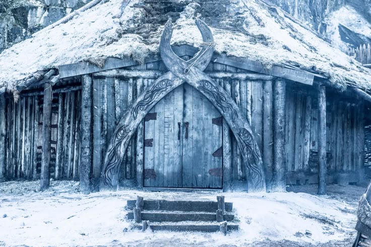 a medium shot shows a wooden building covered in snow during winter. the building features a rustic design with vertically arranged wooden logs. a prominent double door is at the center of the building, adorned with crossed wooden ornaments with engraved designs. a set of three steps lead up to the door. the roof is covered with snow.