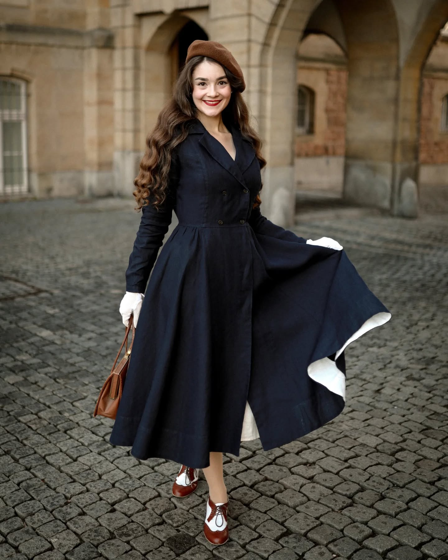 a woman in a navy blue vintagestyle coat dress poses on a cobblestone street.  she is wearing a brown beret, white gloves, and brown and white saddle shoes. she carries a brown handbag. the overall style is reminiscent of the 1940s or 1950s. the setting appears to be an outdoor location with historical architecture in the background. the woman's expression is happy and confident.