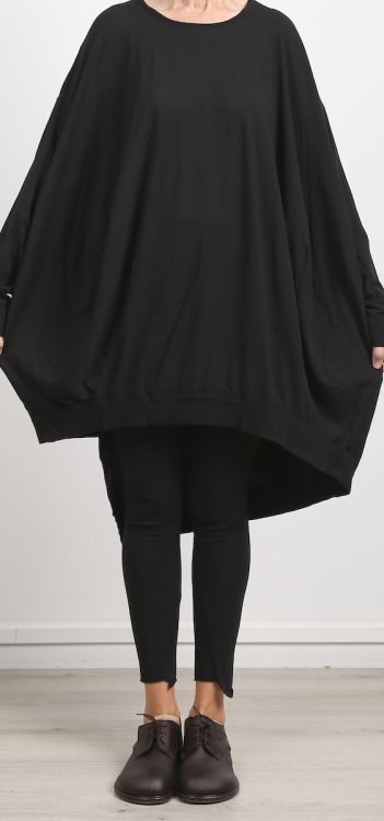 a woman stands facing forward wearing an oversized black top that extends down to her midthighs, paired with black leggings and brown oxford shoes. the clothing has a casual and somewhat baggy appearance, suggesting a focus on comfort and a relaxed style. the background is a plain, light wall, which keeps the focus on the clothing and the person wearing it.