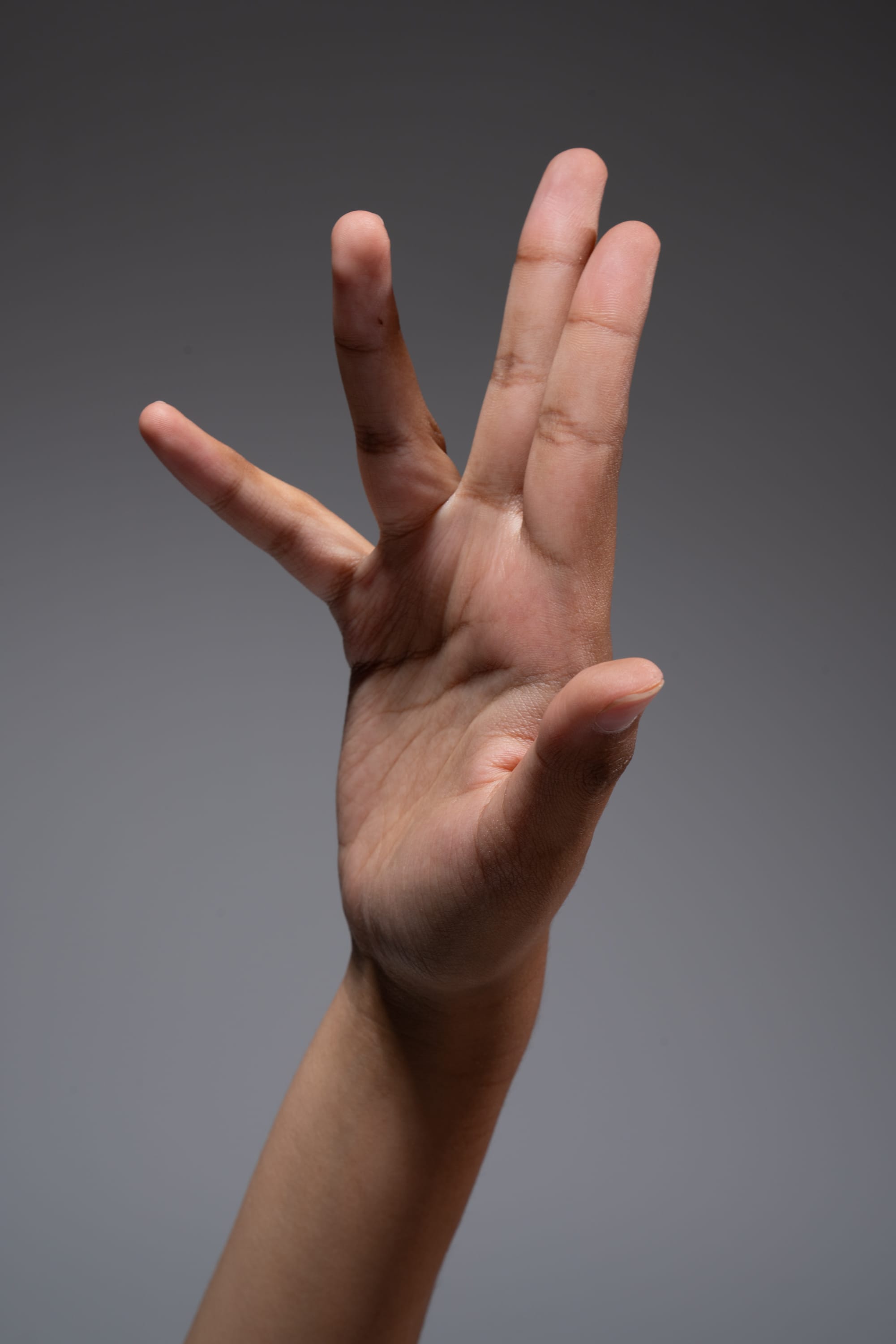 a hand is extended against a gray background, with fingers spread apart in a gesture. the hand belongs to a person with a darker skin tone. the lighting suggests a studio setting, and the closeup perspective focuses attention on the details of the hand.