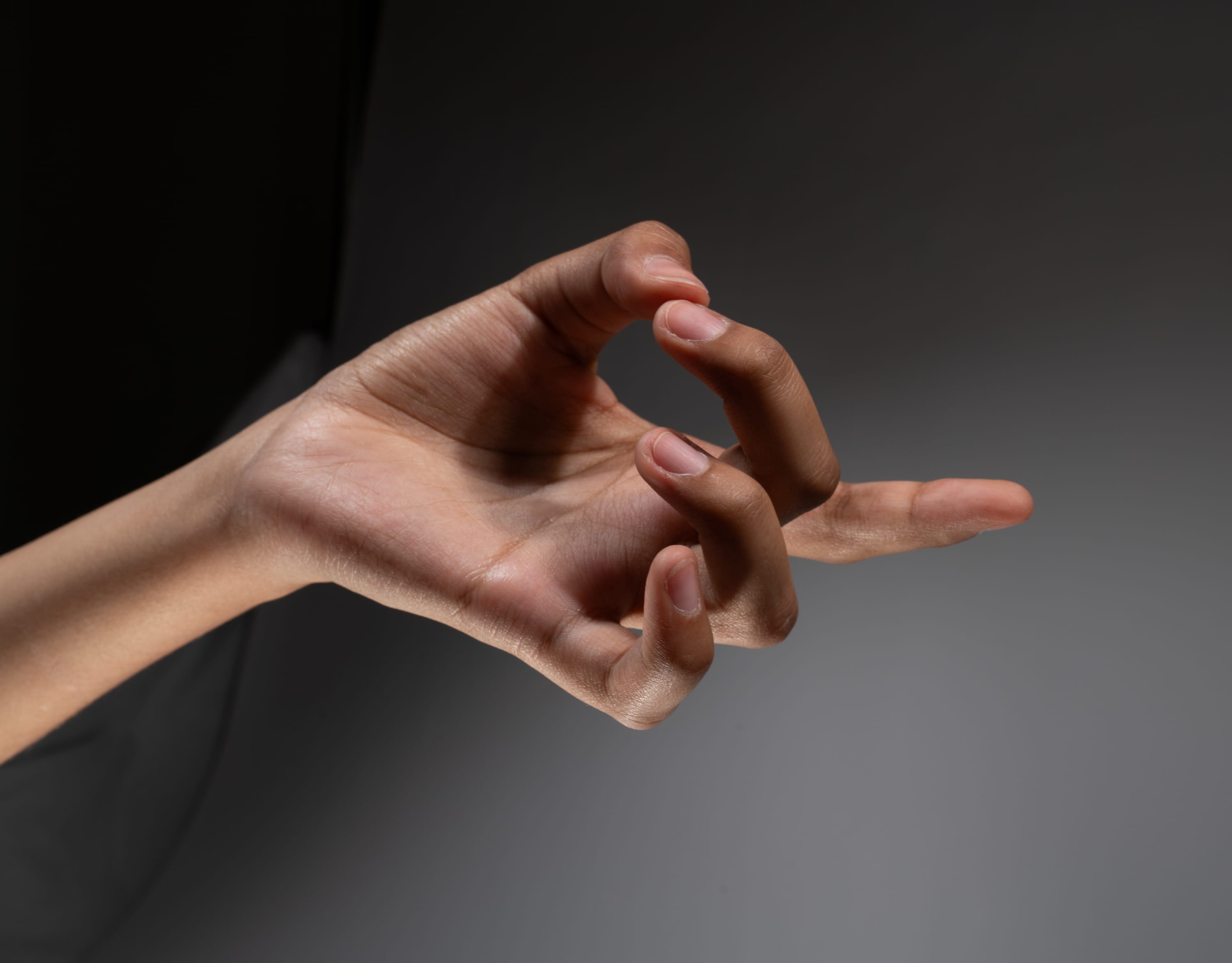 a closeup image of a hand with its thumb and middle finger touching, against a gray and black background.