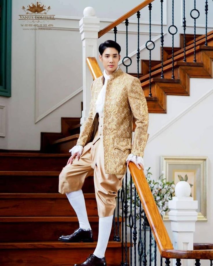 a handsome man in a gold patterned suit stands on a staircase, looking down. he has white socks and shoes, and his suit is accented with white trim. the staircase has a wood handrail and black wrought iron railing. the man is posing for a fashion shoot.