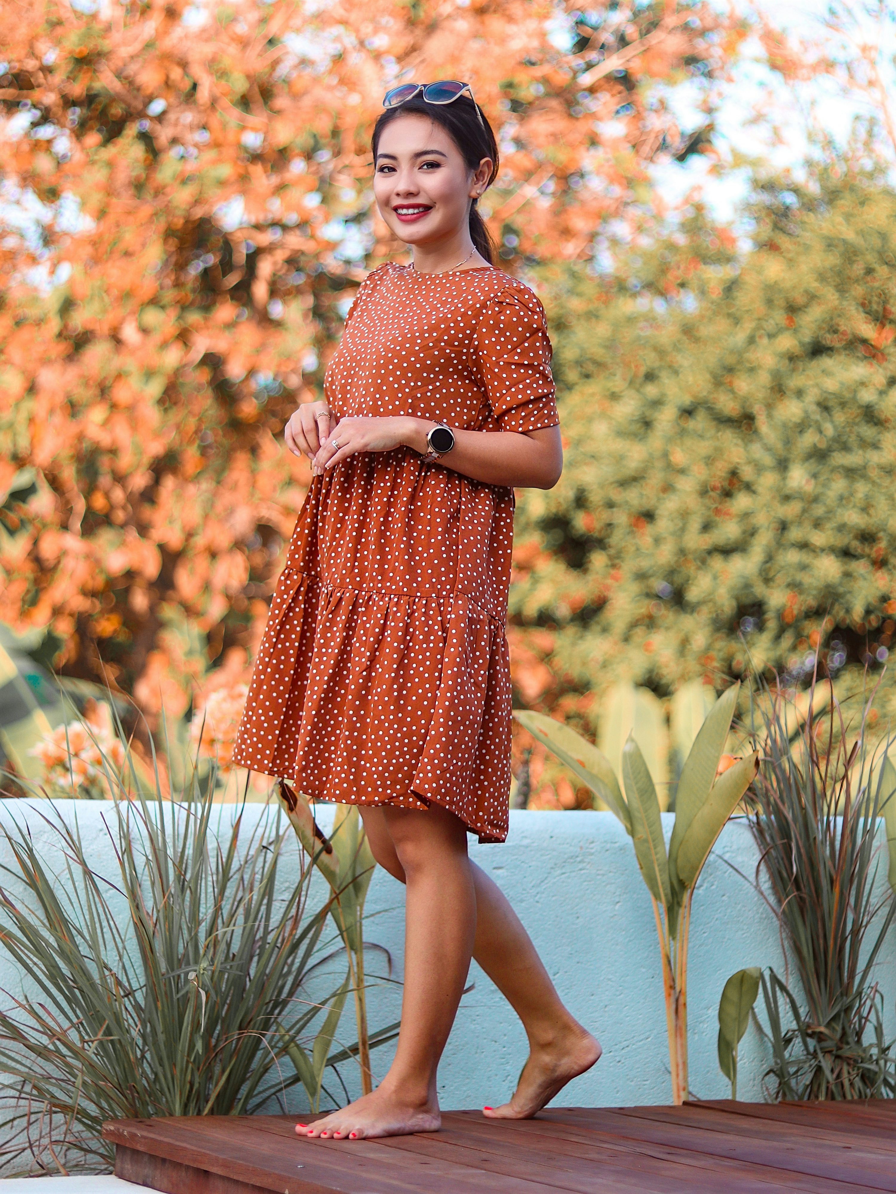 a young woman in a rustcolored polkadot dress stands on a wooden deck outdoors, barefoot and smiling.  the background is blurred but shows autumncolored foliage. the overall mood is bright and cheerful.  the woman is wearing sunglasses and appears relaxed and happy.