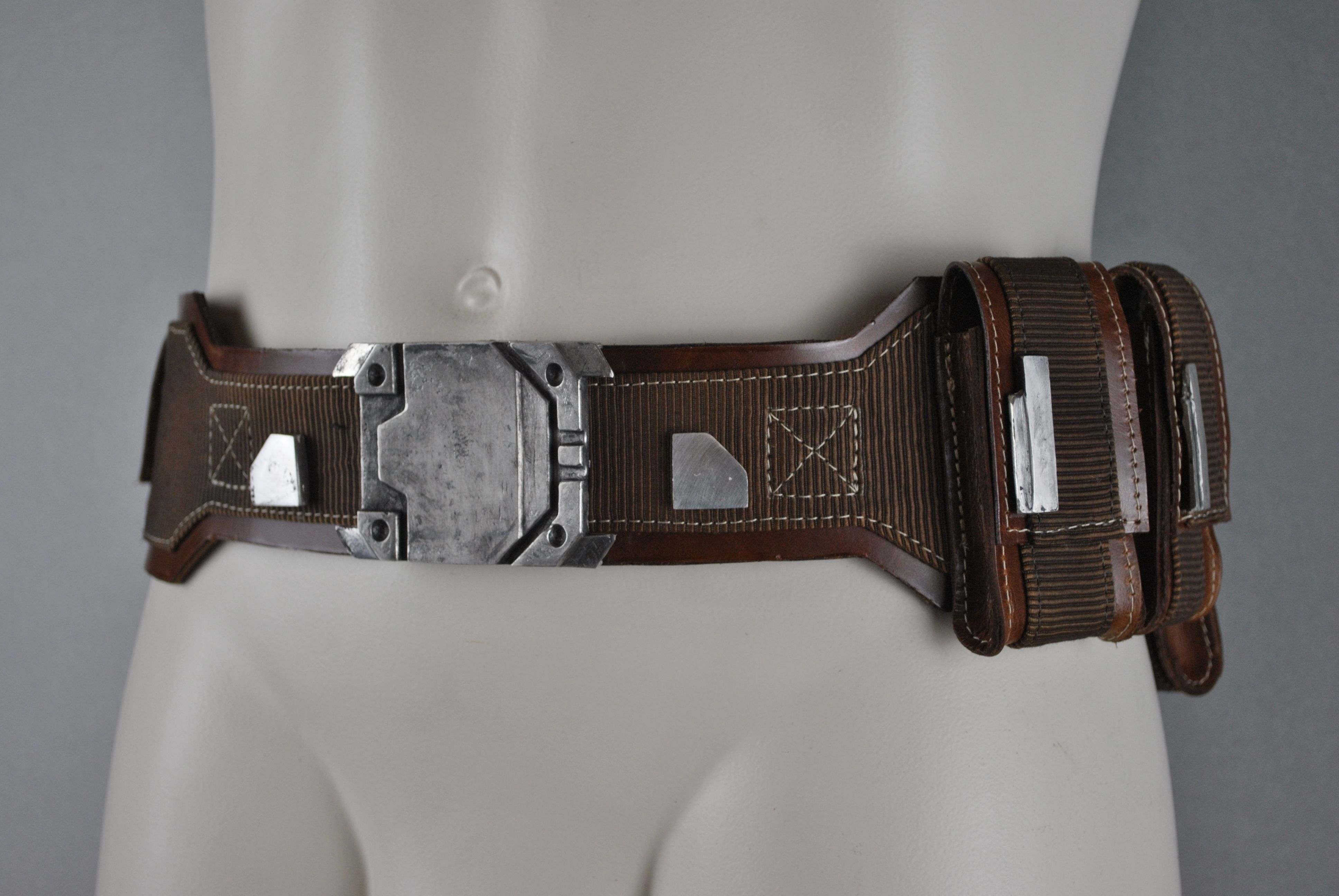 a brown leather belt with a large metal buckle and multiple metal accents. the belt is worn on a mannequin. the belt has a utility style,  with two pouches on the right side.