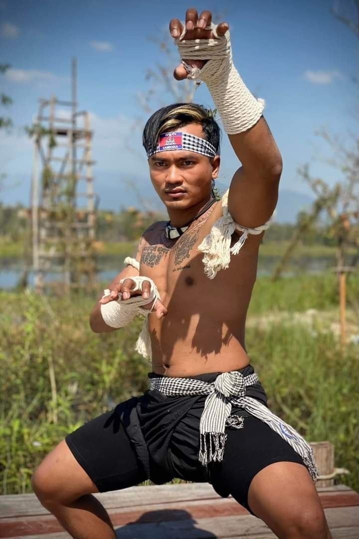 a man wearing traditional khmer muay thai wraps and shorts stands in a fighting pose. he has a headband on his head with the cambodian flag on it and is barechested. he is outdoors in a natural setting with grass and a wooden platform. he is focusing intently on his technique, displaying his strength and determination as a martial artist.
