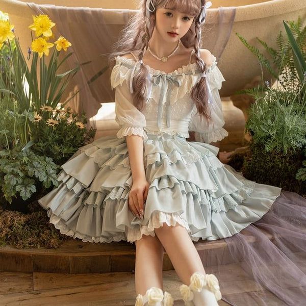 a young woman with blonde hair styled in braids is posed seated on a bench in front of a clawfoot bathtub. she is wearing a light blue and white lolita fashion dress with ruffles and lace detail. she has on white socks with lace and flower embellishments. the image has a soft, dreamy quality and the color palette is light and airy.