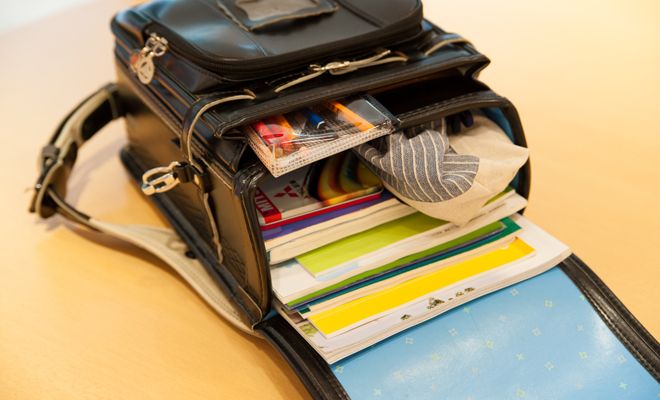the image shows a black leather school bag that is filled with books, notebooks, a pencil case, and a striped gray and white cloth. the bag is open, revealing a light blue interior. silver hardware and buckles are visible on the bag. it is placed on a desk.