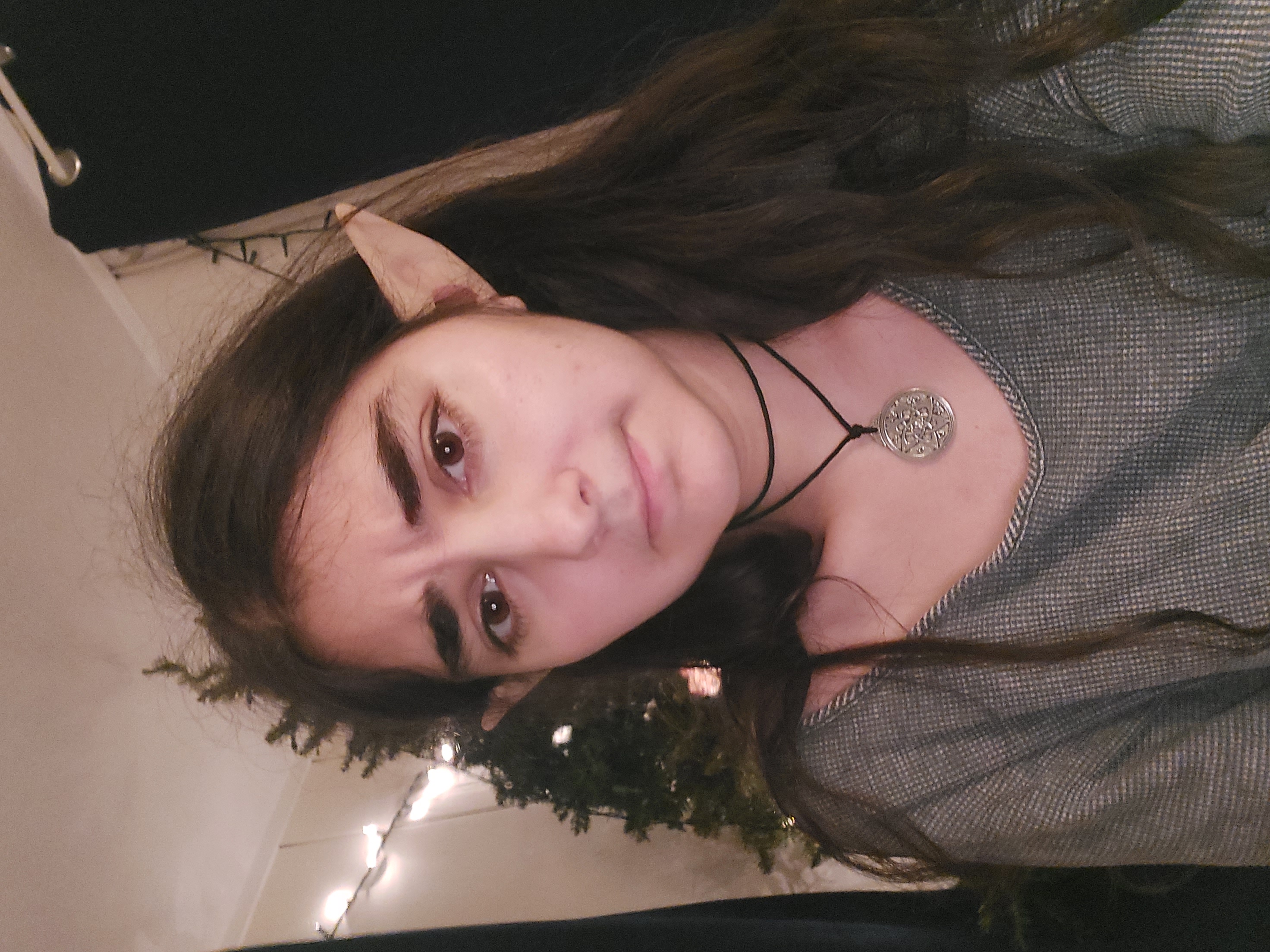closeup selfie of a young woman with long brown hair and elf ears attached. she's wearing a grey sweater, a black choker necklace, and a silver pendant necklace. the background shows a dimly lit room with a christmas tree and string lights. her expression is somewhat serious, almost frowning slightly.  the image has a slightly dark and moody atmosphere.