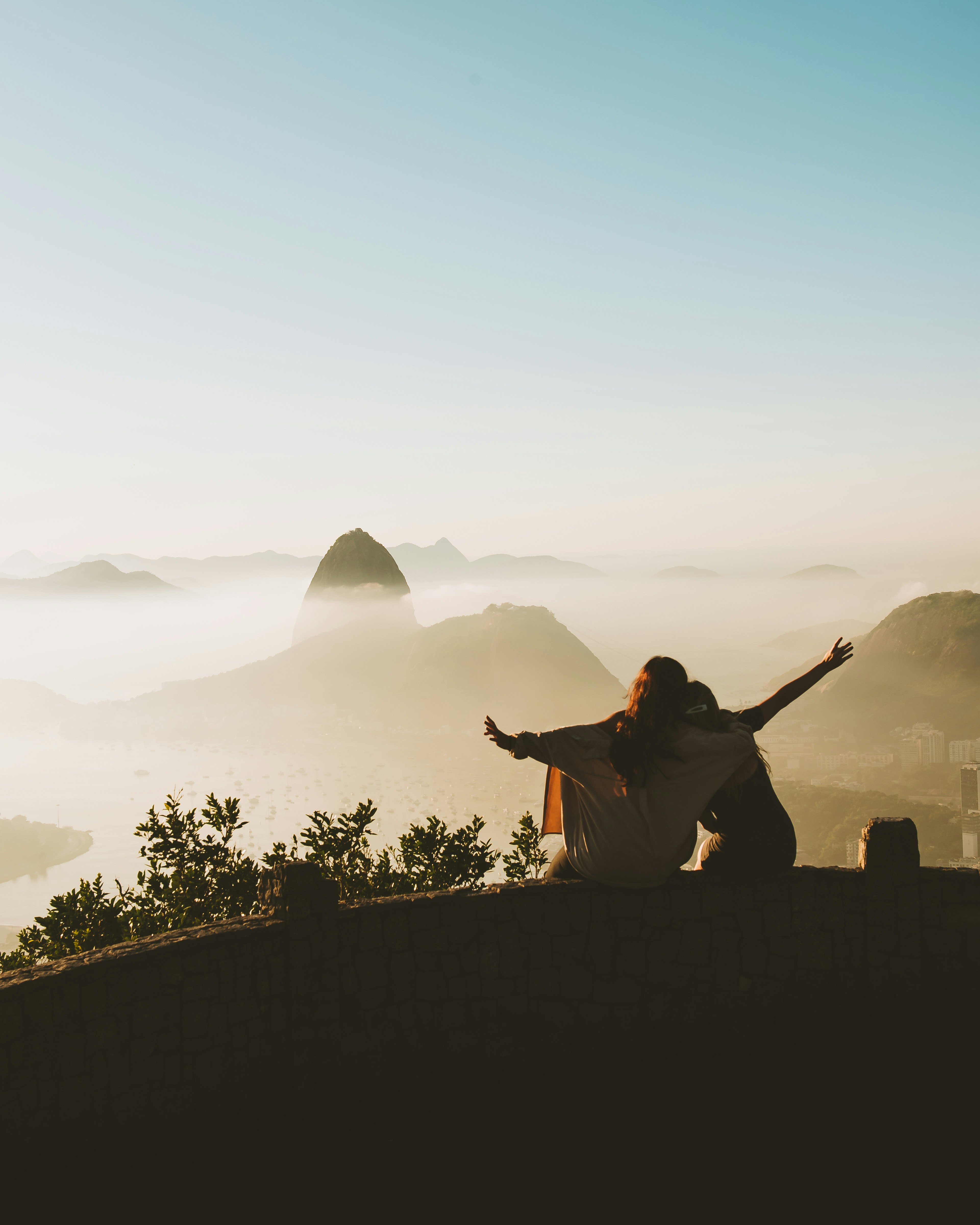 two women, silhouetted against the golden light of sunrise, sit on a wall overlooking a breathtaking vista of rio de janeiro, brazil.  the iconic sugarloaf mountain and christ the redeemer statue are shrouded in mist in the distance as the sun bathes the sky and clouds in warm hues.  the women have their arms outstretched, embracing the beauty and vastness of the scene before them.