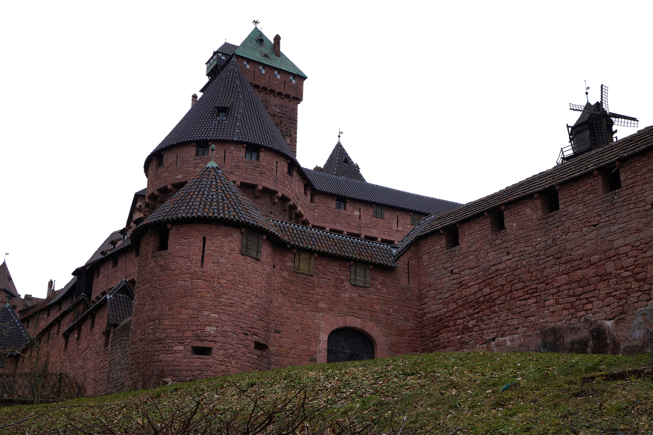 a lowangle shot of a medieval castle made of reddishbrown bricks, featuring towers with conical roofs and a tall central tower with a green roof. the castle is set against a stark black background, and a grassy hill with sparse vegetation is visible at its base.