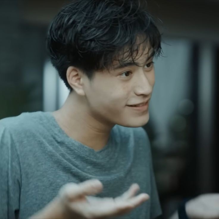 a young asian man is pictured from the shoulders up. he is wearing a bluegrey tshirt.  he is smiling and gesturing with his right hand as though talking to someone offcamera. the lighting is soft, and the background is out of focus.
