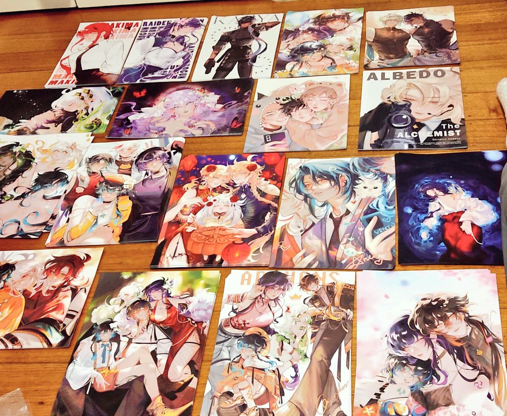 the image shows a collection of animestyle fanart prints, many appearing to be from the game genshin impact.  the prints are laid out on a wooden floor and feature various characters in a variety of poses and settings.  some prints showcase individual characters, while others depict groups of characters interacting.  the art style is consistent across the prints, displaying a vibrant color palette and detailed character designs. the prints appear to be professionally printed and high quality. the overall impression is one of a dedicated fan's collection of artwork.