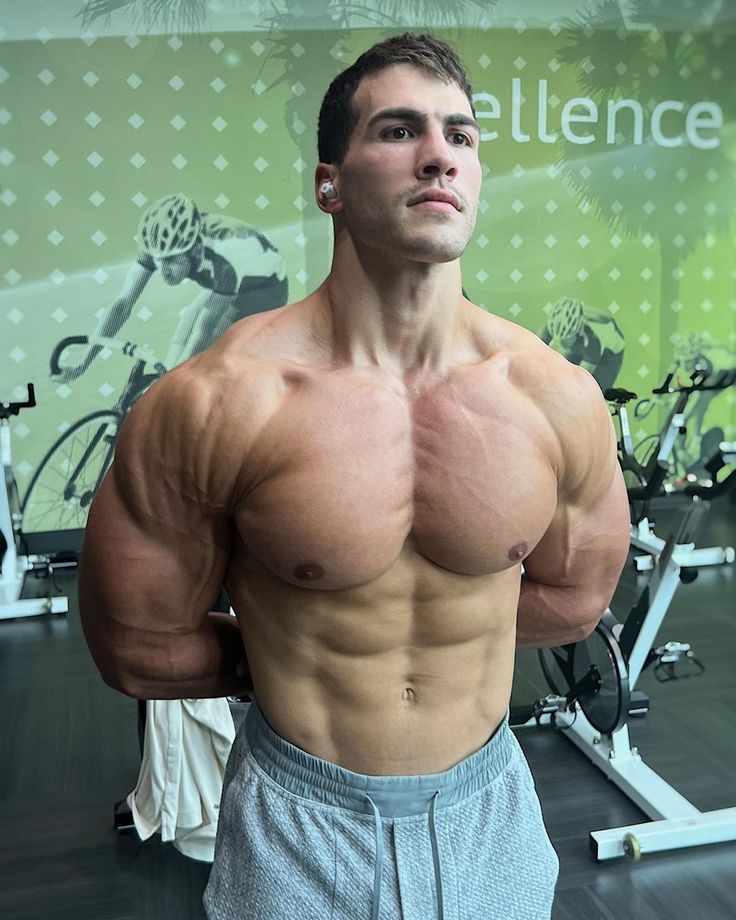 the image shows a muscular man, likely a bodybuilder, standing in a gym. he's shirtless, showcasing a highly developed physique with prominent chest, abs, and arm muscles. he's wearing grey sweatpants and appears to have earbuds in. the background features exercise bikes and a wall with a design incorporating the word "excellence." the overall impression is one of fitness, strength, and physical achievement.