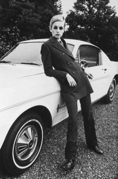 a black and white photo of a woman in a stylish 60s outfit standing next to a white ford mustang. she is wearing a black doublebreasted jacket, pants, and boots. she is leaning against the car, with her hand on the door. she looks stylish and chic. the car is a classic ford mustang, with a distinctive front end and rear lights. the photo is a great example of fashion photography from the 1960s.
