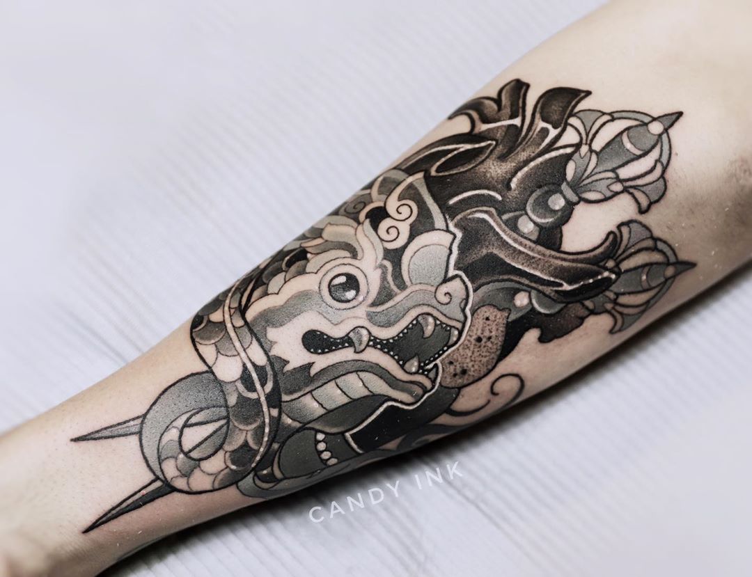 a black and grey tattoo of a dragon head intertwined with a snake is displayed on the inner forearm of a person. the tattoo is done in a neotraditional style with bold lines and smooth shading. the dragon has intricate details and is wearing a headdress. the snake is wrapped around the dragon's head and has its fangs bared. the tattoo is set against the backdrop of the person's fair skin. the lighting is bright, illuminating the tattoo and highlighting the details of the artwork.  the overall impression is one of power, mystique, and artistry.