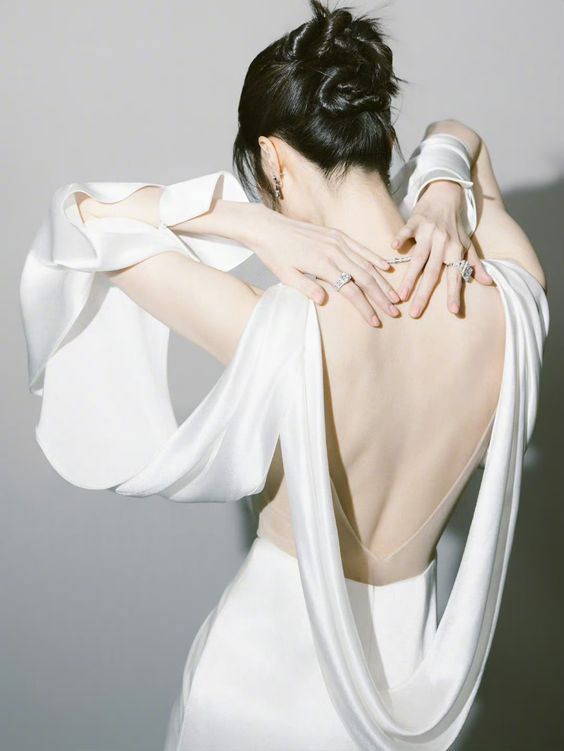 a woman stands with her back to the camera, showcasing the elegant drape of her white silk dress. the dress features long, flowing sleeves and a low, open back.  her hair is styled in a chic updo. the image has a soft and ethereal quality.