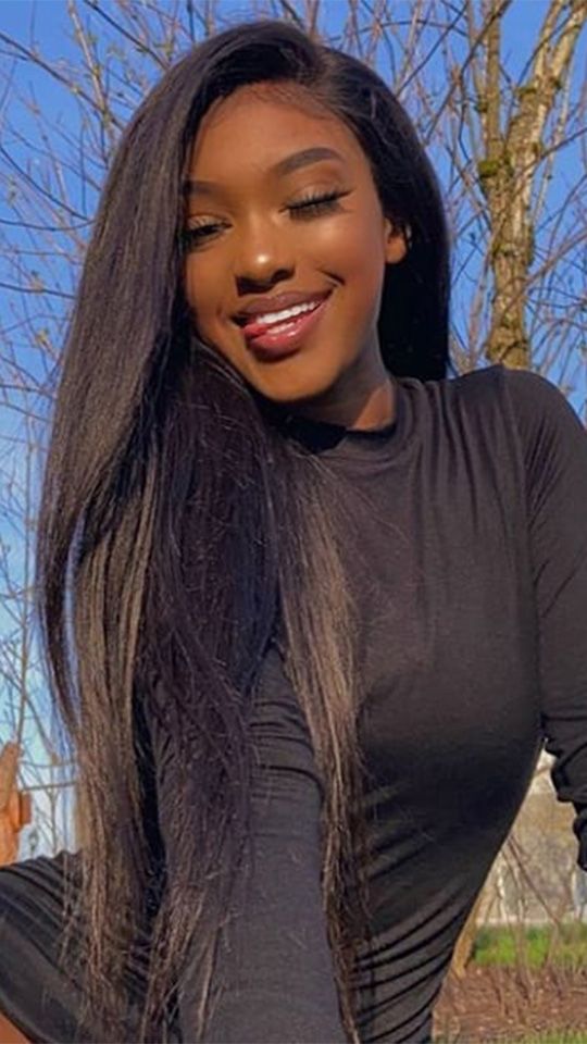 a young woman with long black hair smiles at the camera while sitting on a park bench with a tree behind her. she is wearing a black top and looks happy and confident.