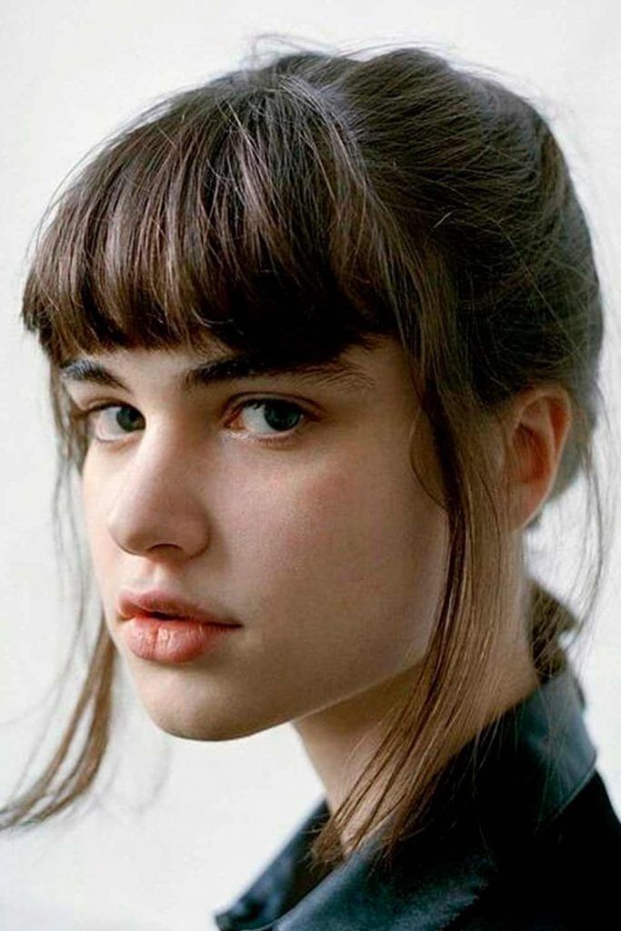 closeup portrait of a young woman with long, straight brown hair and bangs. her hair is parted to the side and styled simply. she has pale skin and a neutral expression. she is wearing a darkcolored collared shirt. the background is plain and white. the overall mood is simple, natural, and slightly melancholic.
