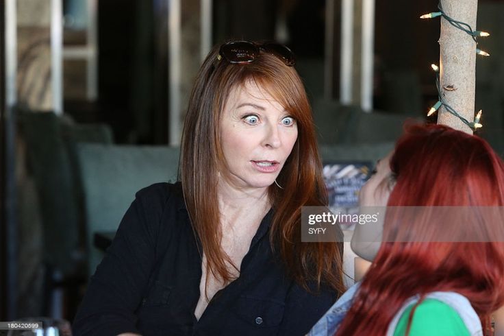 two women sit indoors, one is surprised and looking at the camera, the other is looking to the right. the woman on the left is wearing a black shirt with her reddishbrown hair falling down her shoulders. the woman on the right has bright red hair and is wearing a green shirt and denim jacket.  a string of christmas lights are strung on a tree trunk in the background.