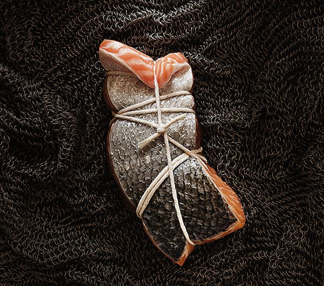the image shows a fillet of raw salmon, carefully tied with twine. the salmon is presented on a dark, textured background that resembles chainmail.  the lighting and composition are artistic, suggestive of a culinary or food photography style emphasizing texture and simplicity. the focus is on the salmon and its presentation; the background is intentionally blurred to keep attention on the subject.