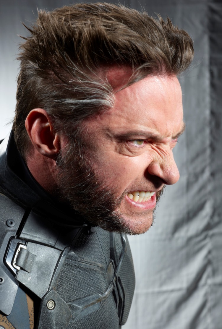 a profile shot of hugh jackman as wolverine, scowling with an angry expression. he has a beard and sideburns with streaks of gray in his hair. he is wearing a black and gray superhero costume.