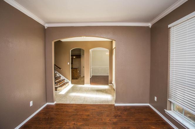 a room with brown walls, hardwood flooring, and a window with blinds. an arched doorway leads to other rooms, including one with tile flooring and a staircase, and another with white wainscoting. the ceiling has white crown molding.