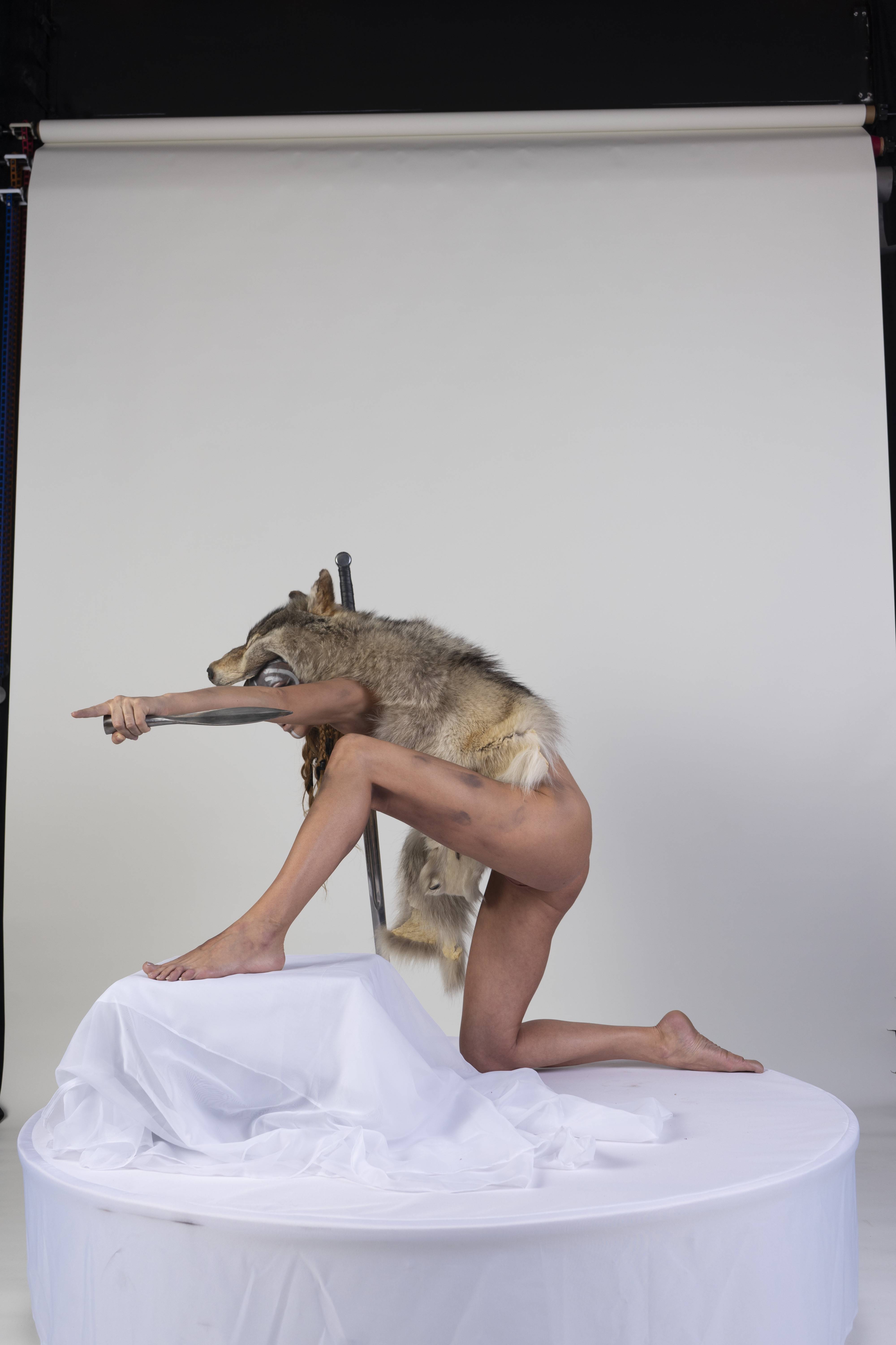 a nude woman kneels on a round, whitedraped stage in a studio setting, against a white background. she is wearing a wolf pelt over her head and shoulders, with the wolf's head covering her face. she holds a metal object, possibly a tool or weapon, in her outstretched hand, pointing forward. her pose suggests a warrior or hunter. the overall image is artistic and surreal, with a raw and primal feel.