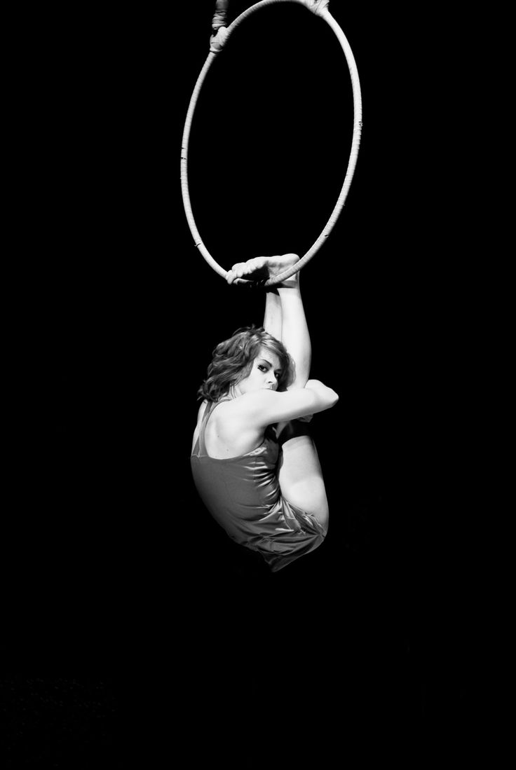 a black and white photo of a woman in a red leotard performing on an aerial hoop. she is suspended upside down and contorting her body, her long hair falling around her. the hoop is a large circle of metal and is silhouetted against a black background. the photo captures the strength and flexibility of the performer.