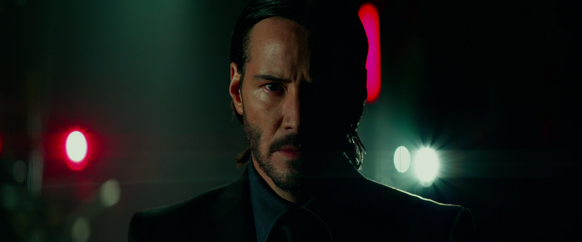 john wick (keanu reeves) in a suit and tie, his face is illuminated by a car's headlights as he stands in a dark street in a city. he's looking directly at the camera. the image is blurry and out of focus in the background.  the image is a still from the movie john wick.