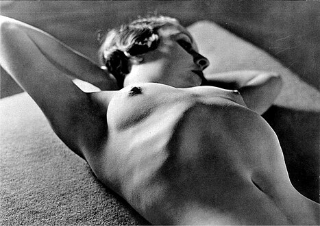the image is a black and white photograph of a reclining nude female figure. the woman is lying on her side, her body angled away from the viewer. her arms are raised above her head, her gaze directed off to the side. the lighting is dramatic, creating strong shadows and highlights that accentuate the curves and texture of her body. the background is blurry and indistinct, drawing all attention to the woman's form. the style of the photography suggests a vintage feel, possibly from the 1920s or 1930s, with a focus on artistic expression of the human form rather than explicit eroticism.