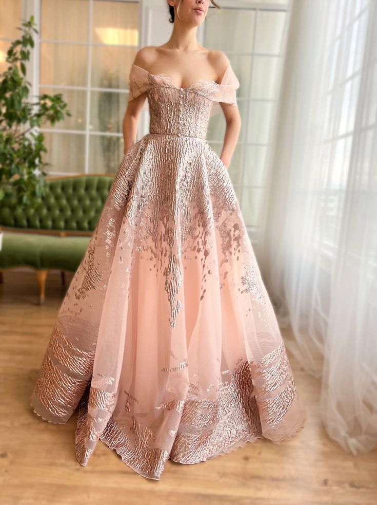a woman in a beautiful off the shoulder dress. the dress is a soft pink with a delicate rose gold embroidery. she is standing in a studio with a white background and curtains in the background. the dress is long and elegant, and the woman looks beautiful.
