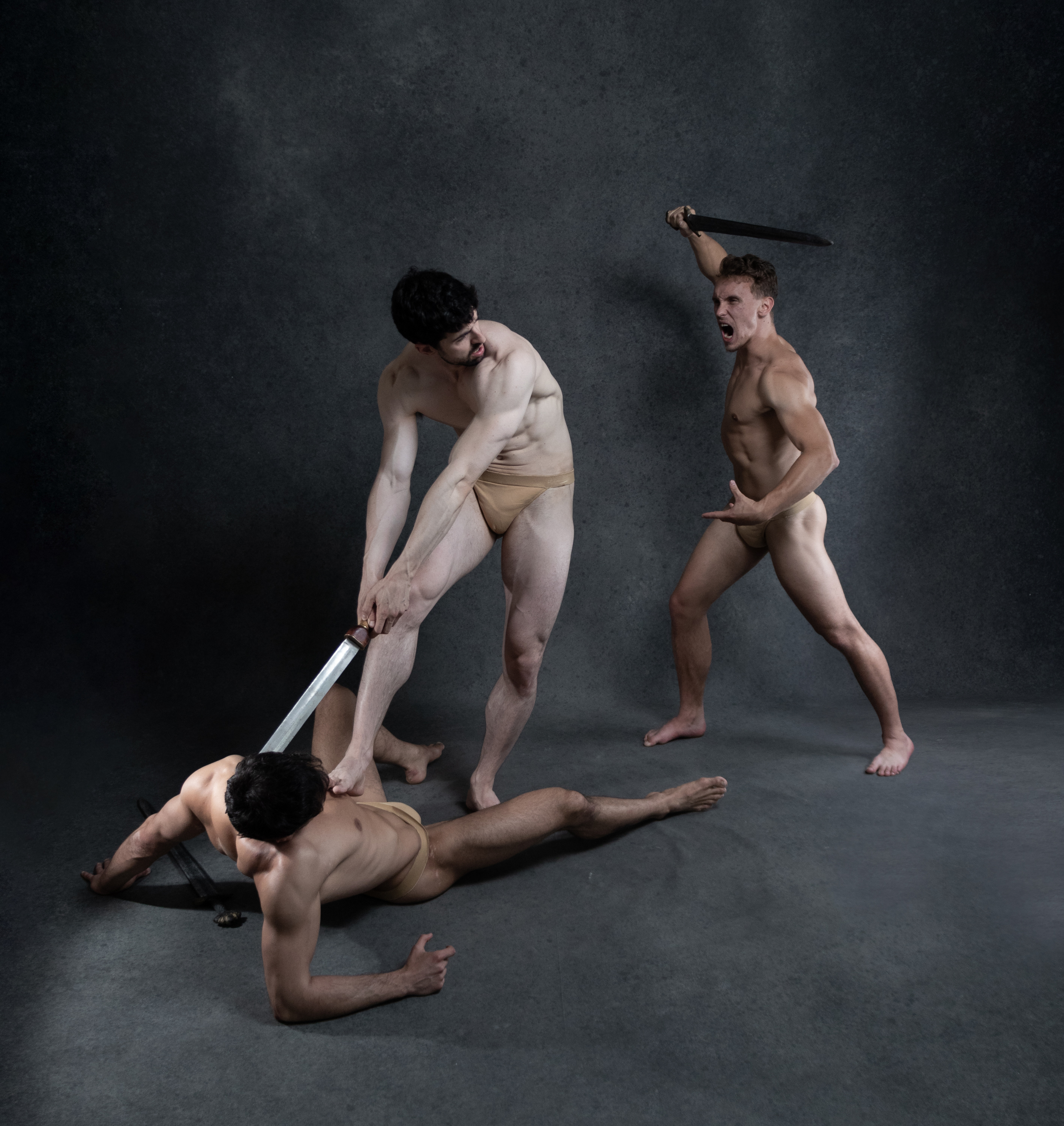 three muscular men are engaged in a fierce sword fight against a dark studio background.  the image is dramatically lit, highlighting the men's physiques and the intensity of their struggle.  one man lies on the ground, seemingly defeated, while another stands triumphantly over him, sword raised.  a third man is caught midlunge, his body a study in dynamic tension. the scene evokes a sense of raw power, aggression, and the primal nature of conflict.