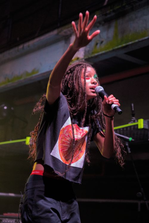 a young woman with dreadlocks sings passionately into a microphone on stage during a concert. she is dressed in a stylish black tshirt with a colorful design and black pants. the stage is dark and the lighting is dramatic, creating an intimate and energetic atmosphere.