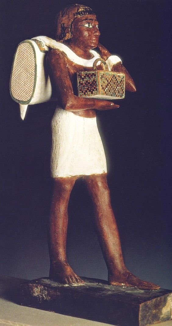 the image showcases a wooden statue of an ancient egyptian man. the statue is depicted in a standing posture, carrying a basket in his left hand and a sack over his right shoulder. the man is adorned with a white kilt, and his skin is painted a deep brown. the basket features intricate, colorful patterns. the background is a dark gray, suggesting the statue is housed in a museum or gallery setting.