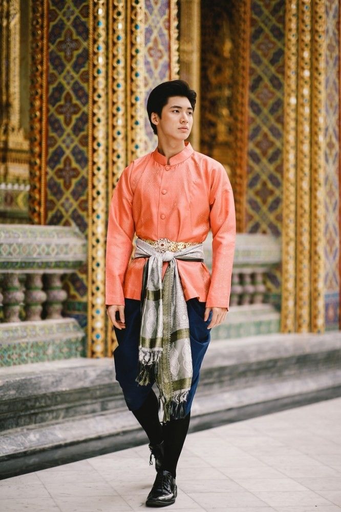 a young man in traditional thai clothing stands in front of a grand ornate building. he is wearing a longsleeved pink shirt and black pants with a colorful sash wrapped around his waist and a matching scarf. his expression is serious, and he looks away from the camera.