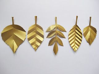 five gold, paper leaves of varying shapes and designs hang against a white background. the leaves are cut out and have a metallic gold finish. their designs include segmented and patterned sections.