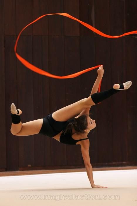 a young woman gymnast performs a ribbon routine, she is holding a red ribbon and is in midair, bending her body in a graceful pose. she is wearing a black leotard and black tights, with her hair pulled back. the background is a wooden floor.