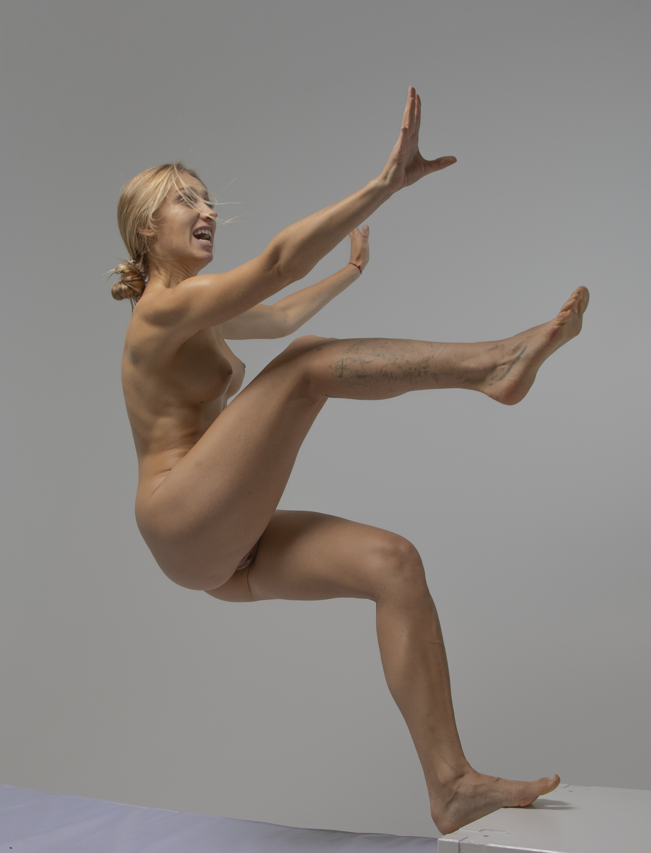 a nude fairskinned woman with blonde hair pulled back is captured midair, appearing to be falling. her arms are raised as if to brace for impact or ward off something, and her mouth is open in an expression of fear or panic. the photograph is a studio shot with a plain gray background. the subject is centered and takes up most of the frame, showing her full body.