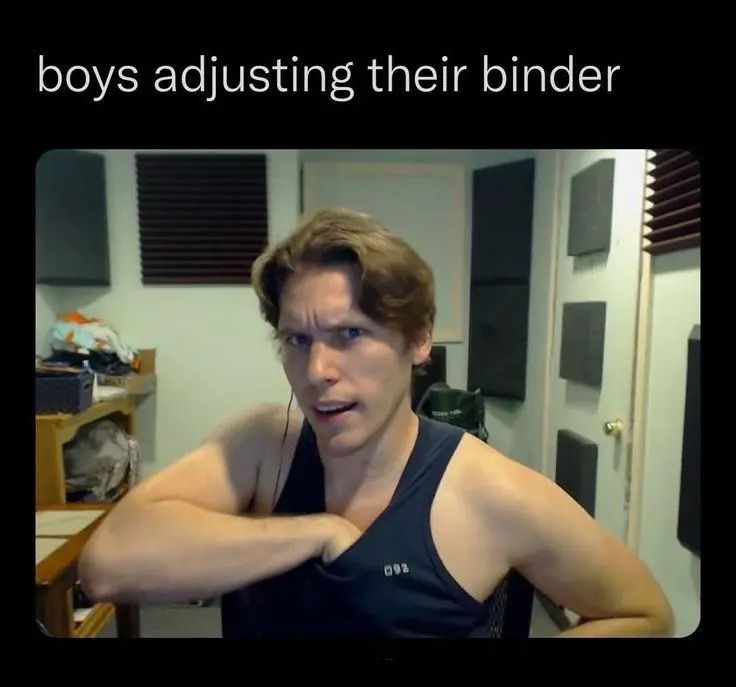 a man with brown hair is seated in a room. he is wearing a tank top and is looking at the camera. he appears to be adjusting something under his shirt.  his expression is serious and he has a microphone attached to his shirt.