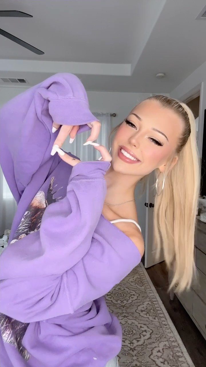 a young woman with blonde hair in a high ponytail is smiling and making a heart shape with her hands. she is wearing large hoop earrings and a purple sweatshirt. she has long nail extensions and is wearing makeup. the background appears to be a bedroom or living room.
