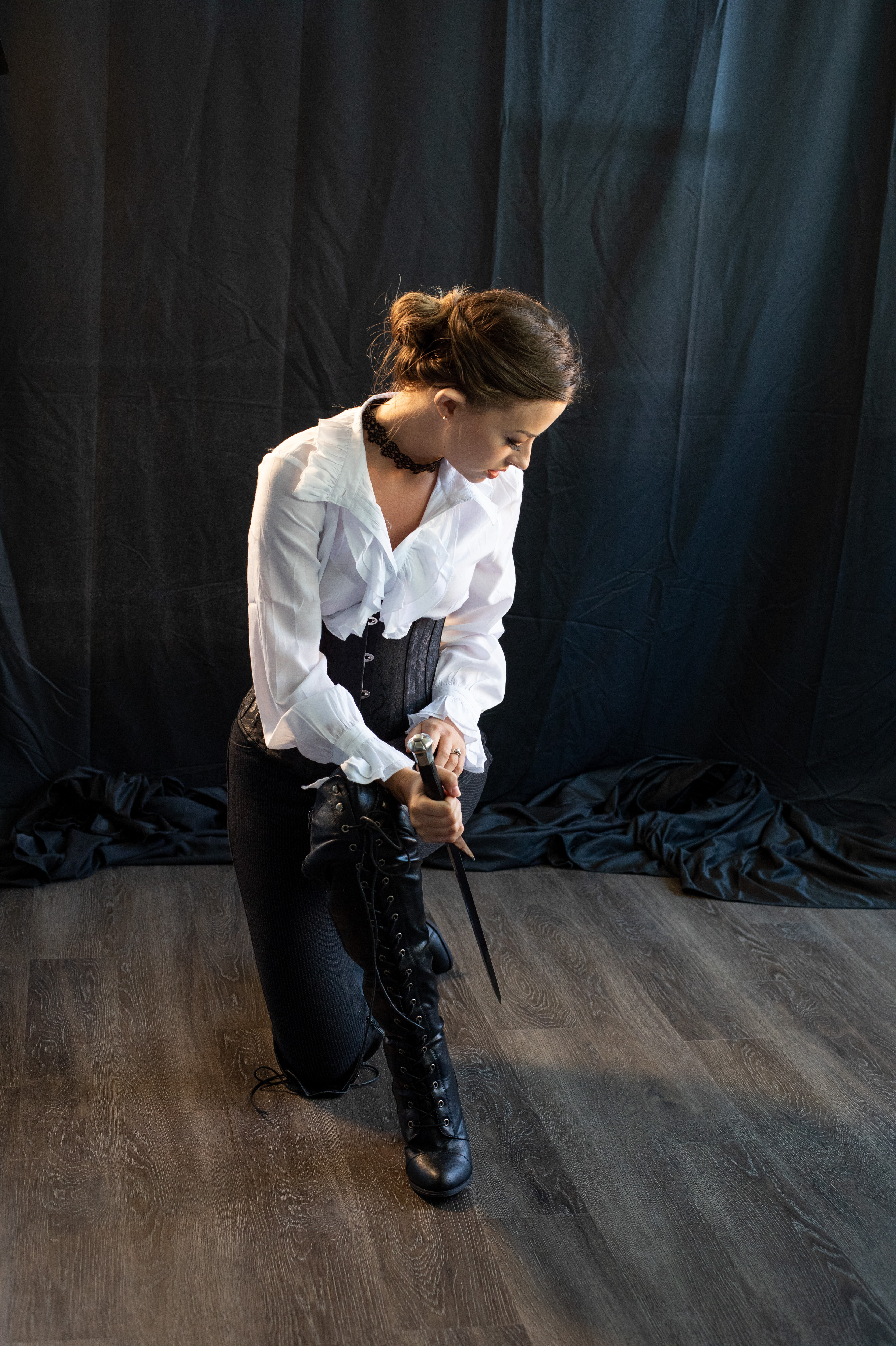 a young woman with her hair pulled back in a bun is dressed in a white, slightly sheer blouse, a black corset, and black pants. she is wearing kneehigh black boots. she is kneeling on one knee on a dark wood floor and holding a sword vertically in front of her, her hands resting on the hilt.  she is looking down at the sword.  the backdrop is dark and textured. the lighting is dramatic and focused on the woman. the overall feel of the image is dark, moody, and strong.