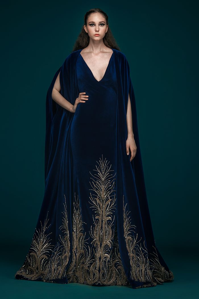 a fulllength studio shot of a model wearing a stunning navy blue velvet gown. the gown features a vneckline and a flowing cape, adorned with intricate gold embroidery that resembles peacock feathers along the bottom hem. the model stands against a teal backdrop, her pose elegant and poised. the image emphasizes the luxurious texture of the velvet and the exquisite detail of the embroidery.