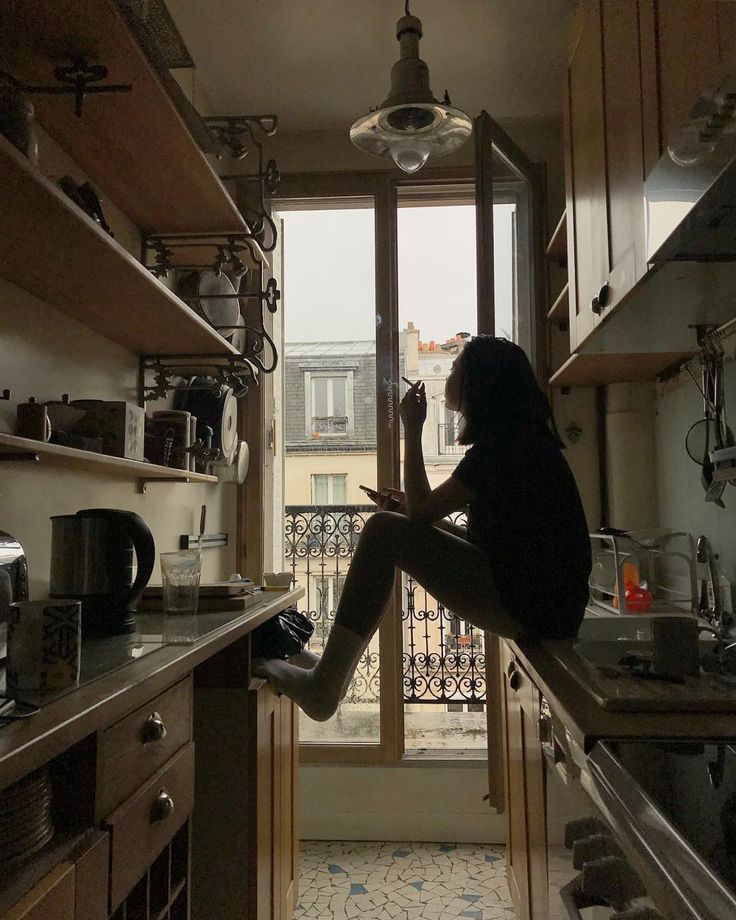 a woman sits on a kitchen counter in a parisian apartment, smoking a cigarette and looking out the window. the kitchen is dark and moody, with vintage wooden cabinets and shelves filled with various items. the window offers a view of the parisian cityscape. the overall atmosphere is cozy and contemplative.