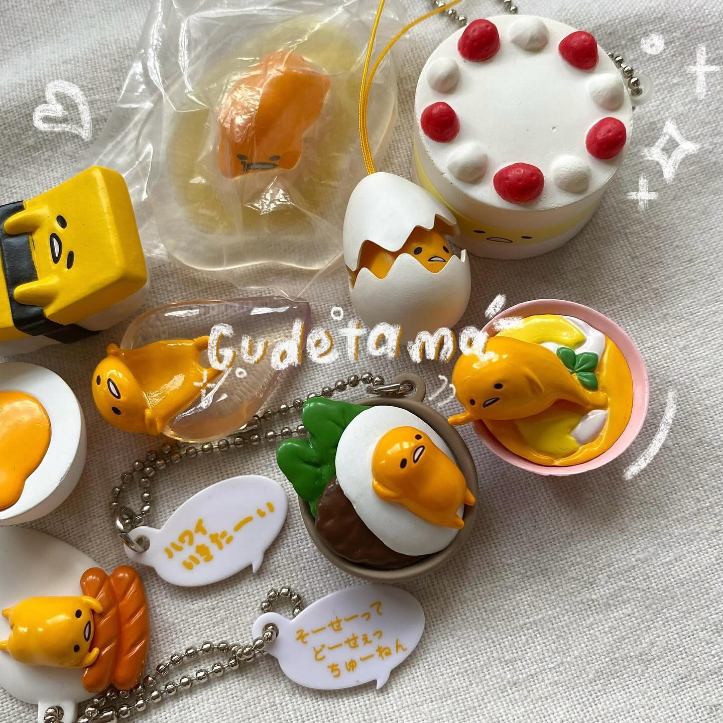 the image displays a collection of gudetama themed keychains and charms arranged on a textured surface. there is a variety of designs, including gudetama in an eggshell, on a cake, in soup, on a hamburger, in a plastic bag, and laying on a hotdog. each keychain has a corresponding tag with japanese writing on it.