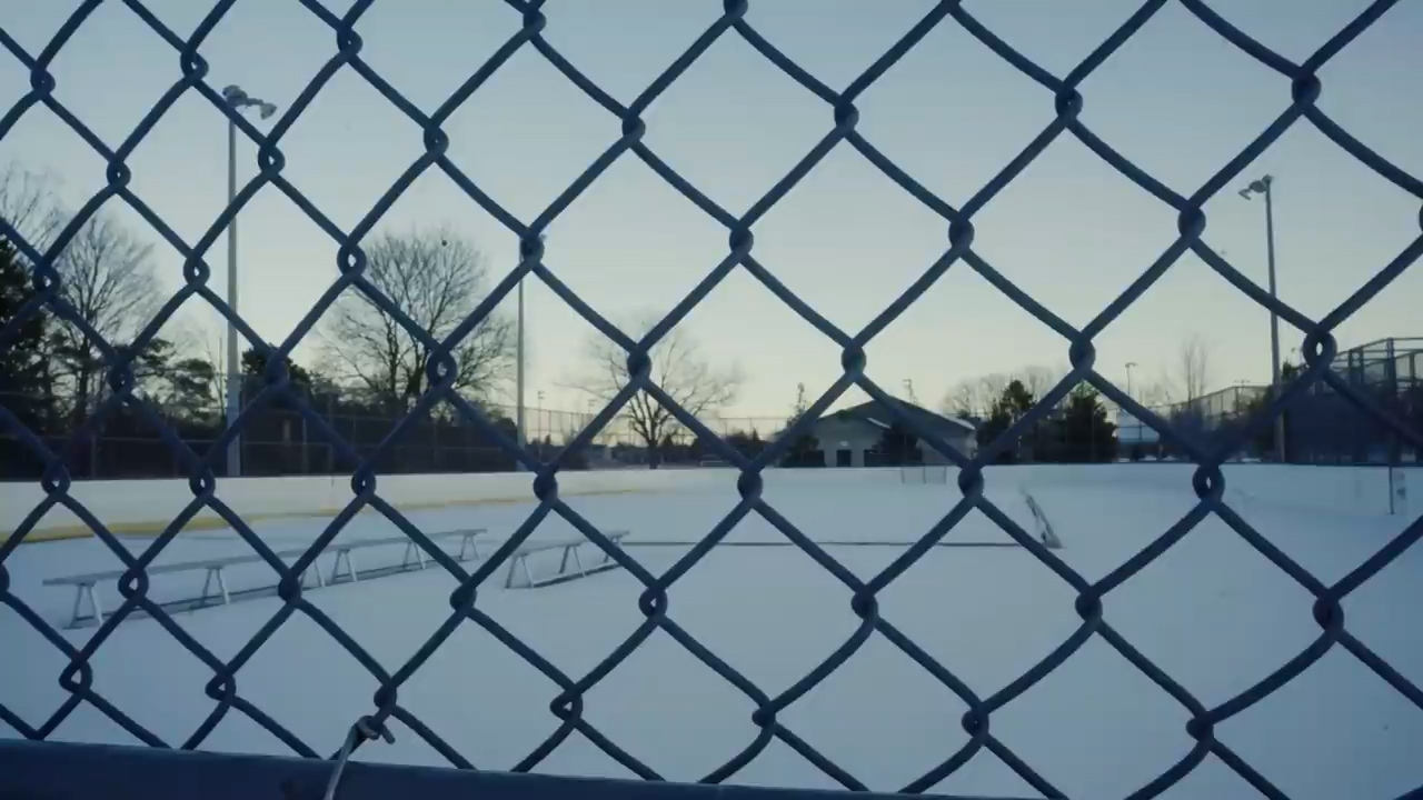 the image shows a snowcovered outdoor hockey rink viewed through a chainlink fence. the rink is surrounded by trees and a fence. the sky is a pale blue, and the snow is white. there are benches on the left side of the rink. the overall tone of the image is cold and serene.