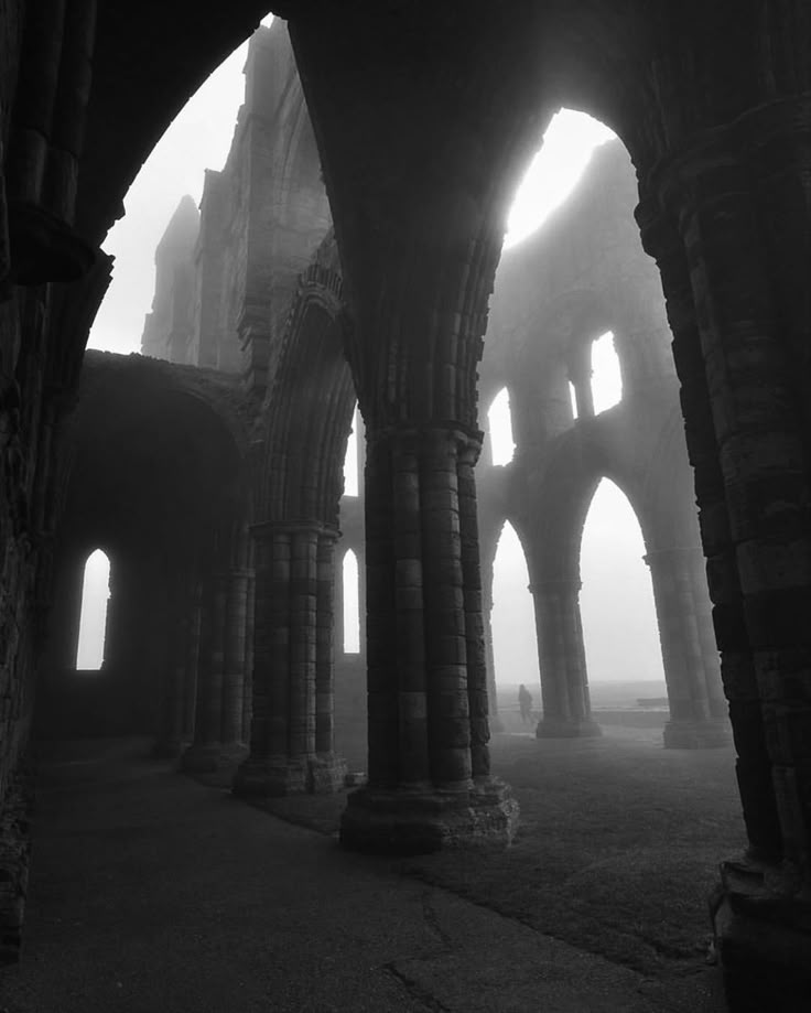 a black and white photo shows the ruins of whitby abbey, featuring stone arches, columns, and a foggy atmosphere. a pathway and grass are visible, and a silhouette of a person can be seen in the distance. the image conveys a sense of history, age, and mystery.