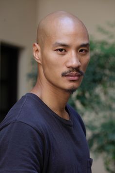 a bald asian man with a serious expression, wearing a dark blue tshirt, stands in an outdoor setting with a blurred green background. the lighting is natural and the perspective is at eye level.