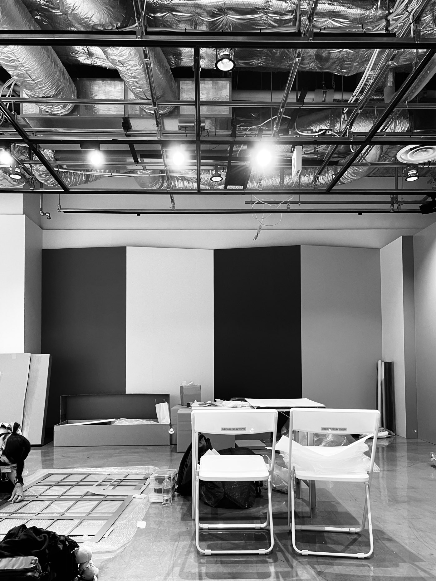 the black and white image shows a room under construction or renovation. there are exposed ceilings with ductwork and lighting fixtures. the walls are partially painted in a geometric pattern of white and gray. two white folding chairs and a table are placed in the center of the room.  construction materials are scattered around, suggesting ongoing work.  the overall impression is one of incompleteness, with the stark black and white emphasizing the industrial feel of the space.