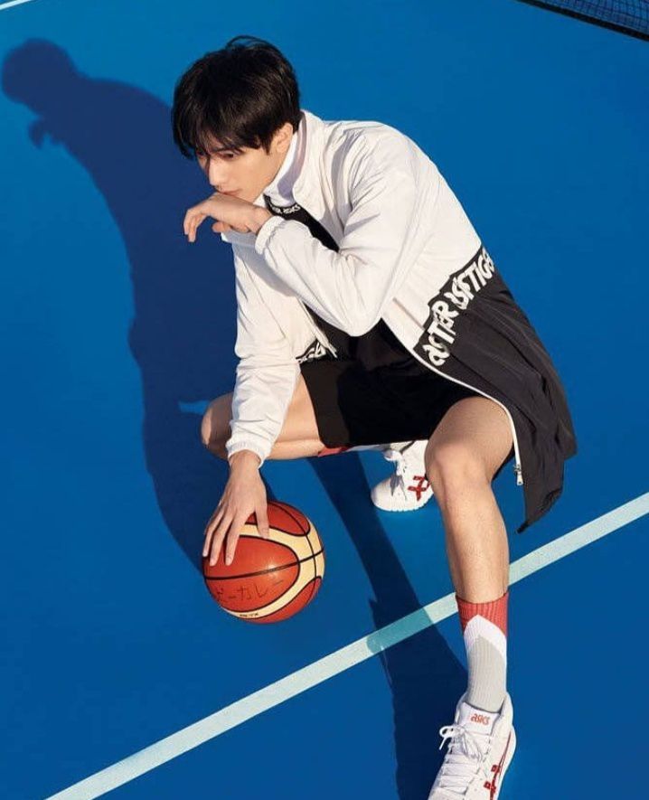 a young man is crouching on an outdoor basketball court, holding a basketball. he's wearing a white jacket with black details, black shorts, white socks with a red stripe, and white sneakers. the court is blue, and the lines are white. the photo is taken from a high angle, and the lighting is natural and bright.